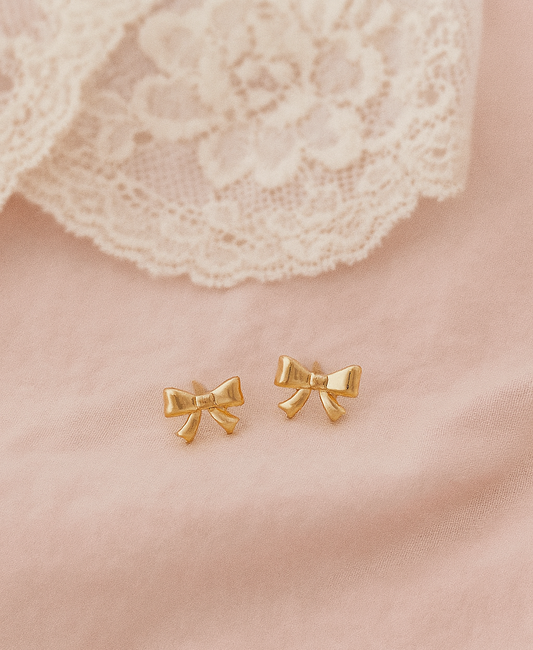 Gold bow-shaped earrings on a soft pink fabric background