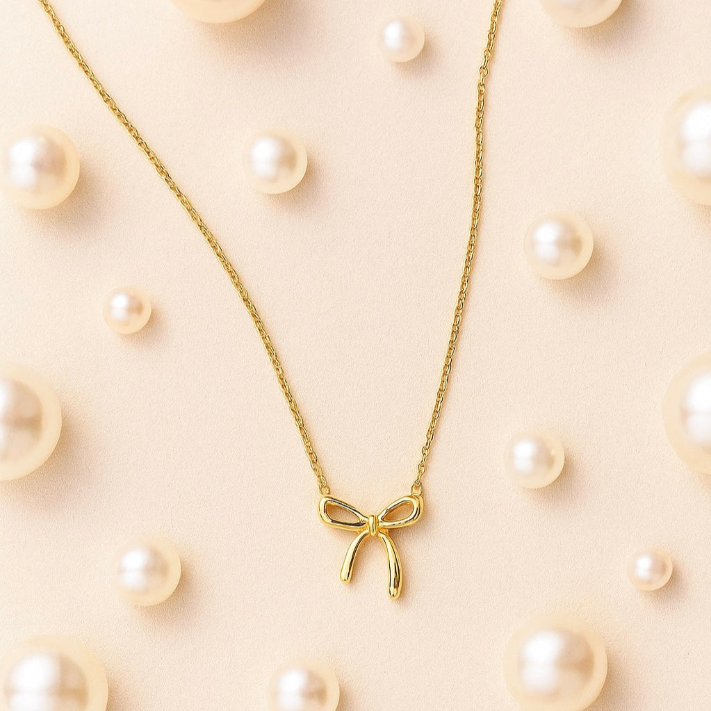 Gold bow necklace on a beige background with scattered pearls